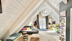 A cozy attic bedroom with a sloped ceiling, a large window, a bed, a desk, and a bookshelf.