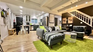 A modern living room with a grey sofa, a coffee table, and a green rug. There is a kitchen in the background with white cabinets and a stainless steel refrigerator.