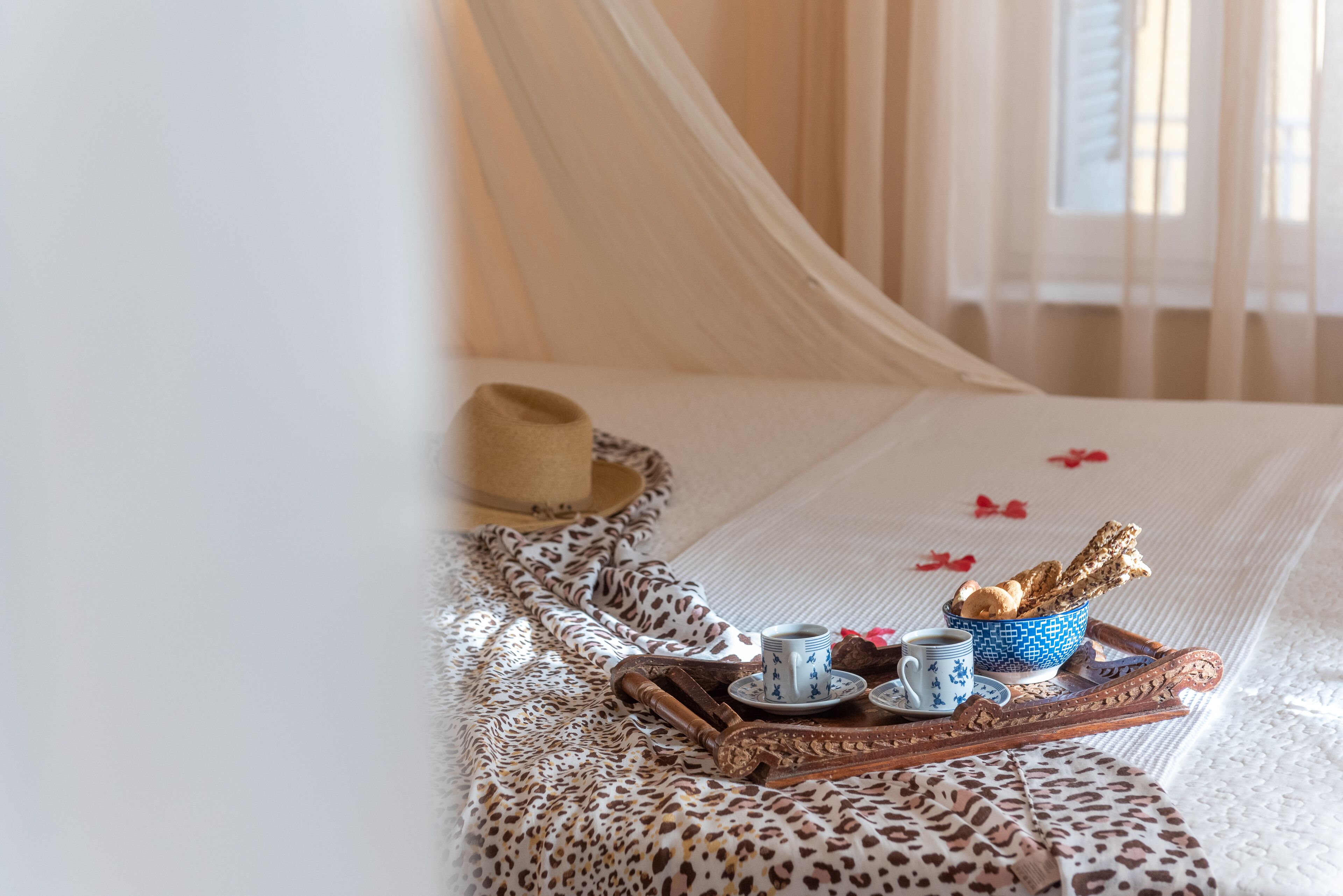 Ein Bett mit weißer Bettdecke, einer Decke mit Leopardenmuster, einem Holztablett mit Tassen und Gebäck sowie einem Hut auf dem Bett.