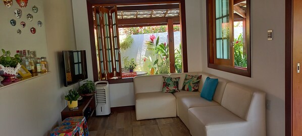 A cozy living room with a white sofa, a small table with a colorful tablecloth, a heater, and a window with a view of a garden.