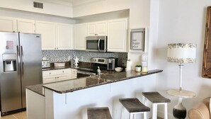 Una cocina moderna con armarios blancos, un refrigerador de acero inoxidable, un microondas y una encimera de granito. Hay tres taburetes de madera en la isla.