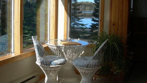 A cozy room with a wooden ceiling and large windows overlooking a lake. There is a table with two wicker chairs and a potted plant.