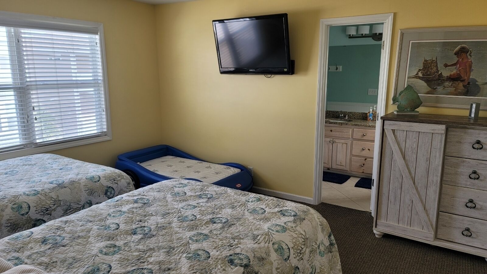 Une chambre avec deux lits, un téléviseur fixé au mur, une commode et une salle de bain visible à travers une porte ouverte.