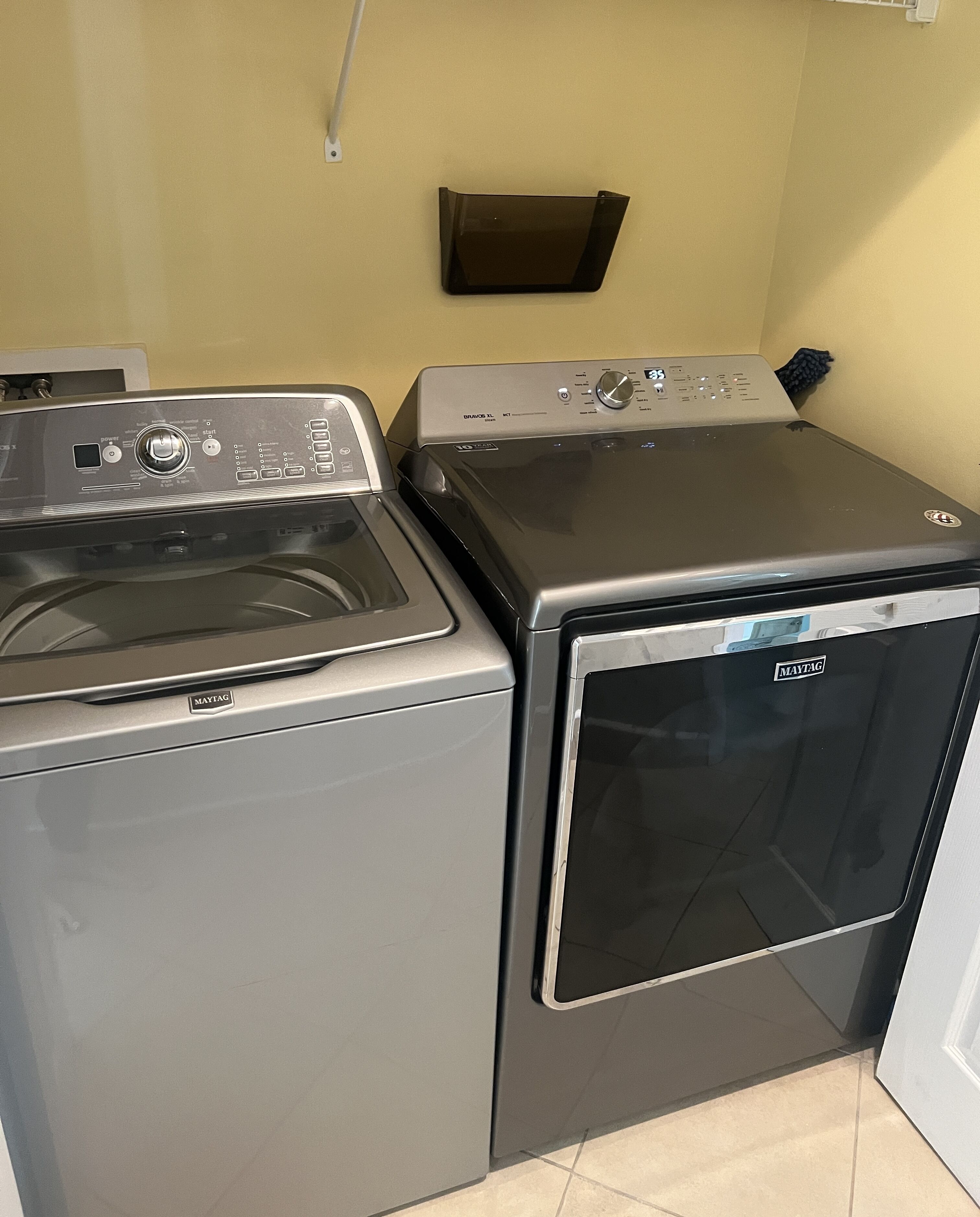 Een kleine wasruimte met een Maytag wasmachine en droger met voorlader, een klein plankje boven de droger, en een betegelde vloer.