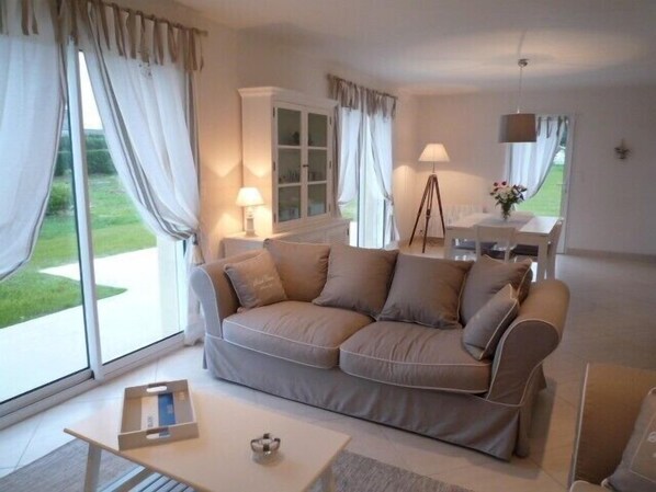 A cozy living room with a beige sofa, a small table, and a lamp. There is a window with curtains and a view of greenery outside.