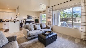 A spacious living area with a grey sofa, a black ottoman, and a dining table. A kitchen with white cabinets and a central island is visible in the background. - Grayhawk Mountain Views! Highly desirable Tesoro unit! (Scottsdale)