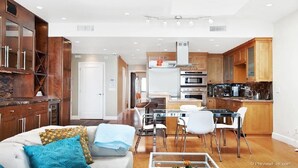 Una cocina moderna con armarios de madera, electrodomésticos de acero inoxidable y un comedor con mesa de vidrio y sillas blancas.