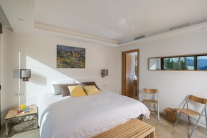 A modern bedroom with a large bed, a painting on the wall, a bedside table with a lamp, and a view through a window.
