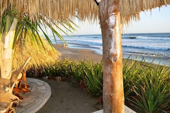 A beach scene with a thatched roof structure, palm trees, and a sandy path leading to the ocean.