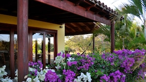 A covered outdoor area with a wooden pergola, surrounded by lush purple and white flowers, and a view of a tropical landscape. - Relaxing Beach House Just Steps from Nicaragua's Best Surf Breaks (San Diego)