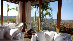 Ein Zimmer mit einem weißen Sofa und einem Sessel, einem Holztisch mit einer Schale und Blick auf eine üppige Landschaft mit Palmen und Bergen.