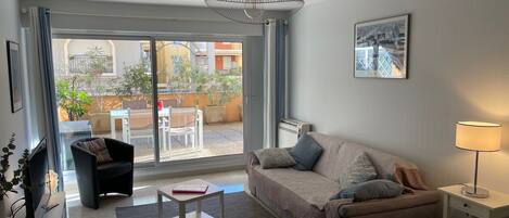A modern living room with a sofa, a coffee table, a TV, and a dining area visible through the glass door.