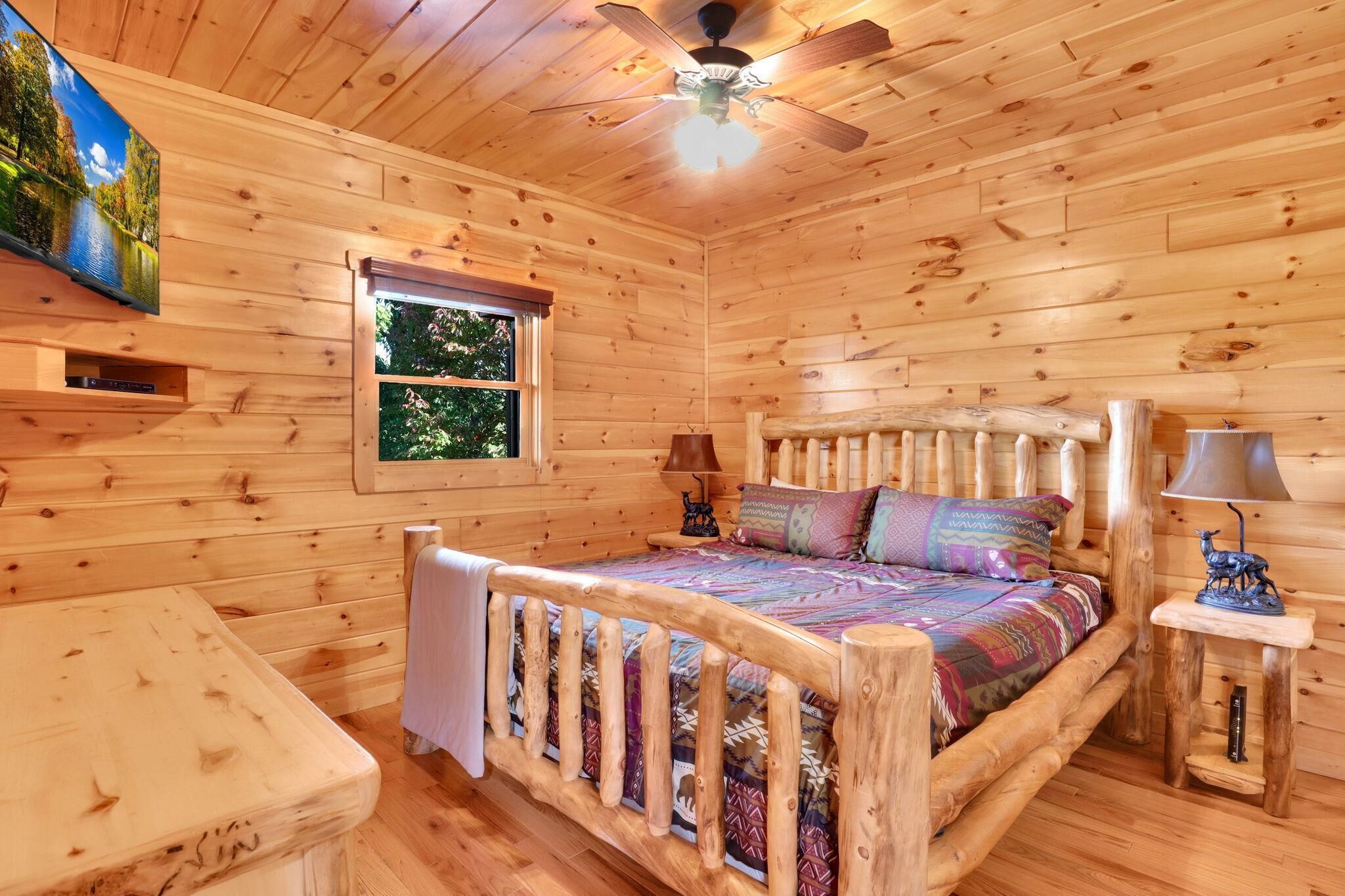 A wooden cabin bedroom with a bed, a ceiling fan, a window, and a TV mounted on the wall.