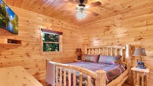 Un dormitorio tipo cabaña de madera con una cama, un ventilador de techo, una ventana y un televisor montado en la pared.