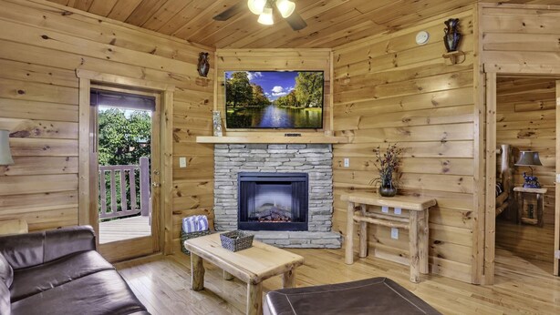 Interior acogedor de una cabaña de troncos con chimenea de piedra, un televisor de pantalla plana montado encima, sofás de cuero y un ventilador de techo de madera.