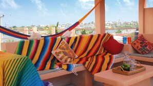A hammock on a balcony with colorful cushions, a small table with a tray of fruit and drinks, and a view of a cityscape.