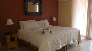 A neatly made bed with white linens, two bedside lamps, a wooden nightstand, a mirror on the wall, and an air conditioning unit.