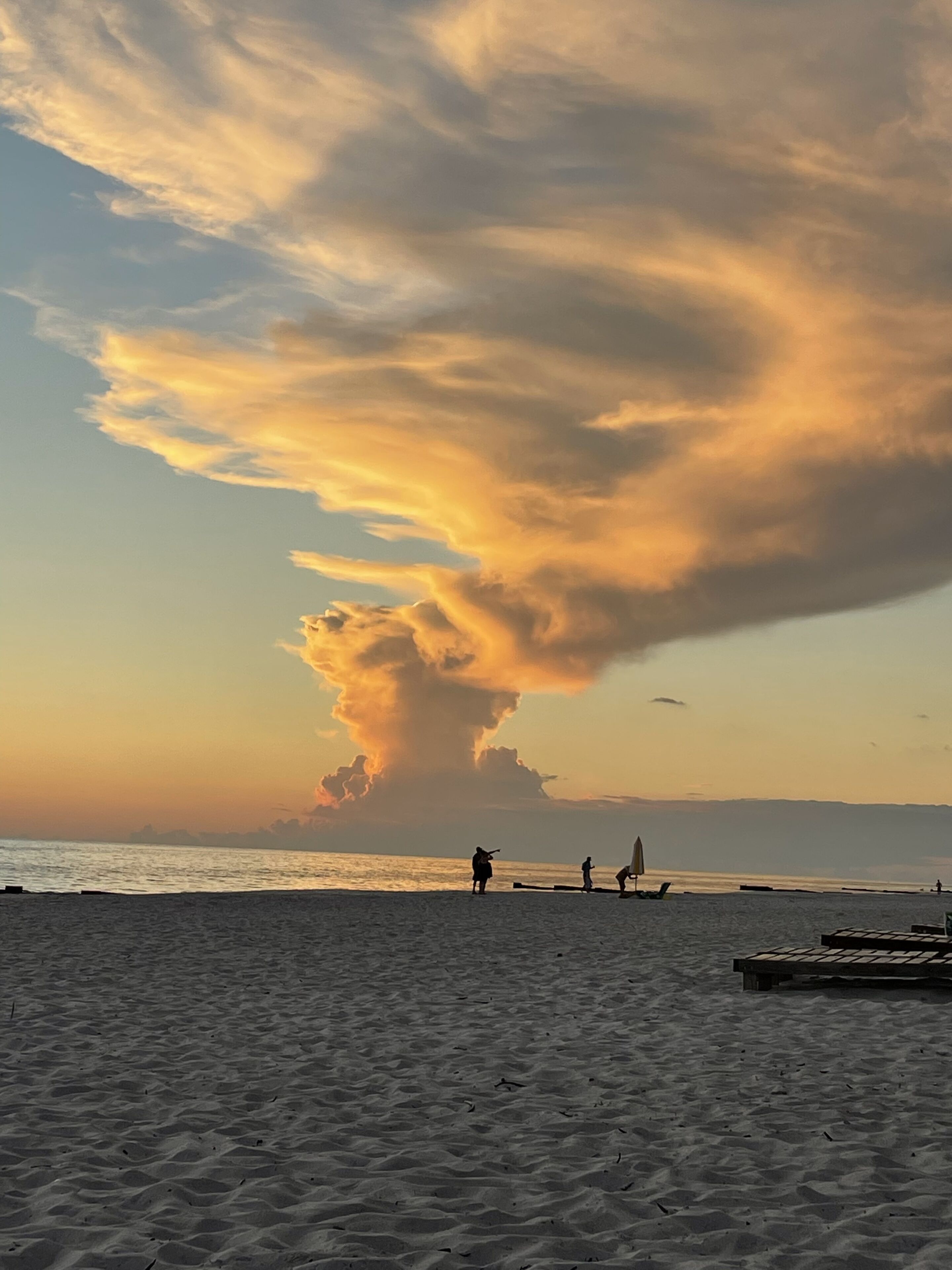Una playa con una gran formación de nubes en el cielo, gente caminando y una estructura de madera a lo lejos.