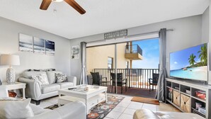 Un salon moderne avec un canapé blanc, une table basse et un téléviseur à écran plat. Il y a un balcon avec vue sur l’océan et une pancarte sur laquelle on peut lire : « Le paradis est un peu plus proche : dans une maison au bord de l’eau. »