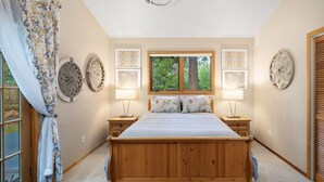 Une chambre à coucher avec un lit en bois, deux tables de chevet avec des lampes, une fenêtre donnant sur un paysage verdoyant et des œuvres d’art décoratives accrochées au mur.
