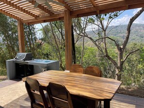 An outdoor patio with a wooden table and chairs, a grill, and a view of a mountainous landscape.