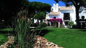A white building with a tiled roof and a small tower, surrounded by green grass and trees. - Luxury villa V2, sleeps 6, Elevator to the beach, Cliff, Pine Cliffs Golf (Olhos de Agua)