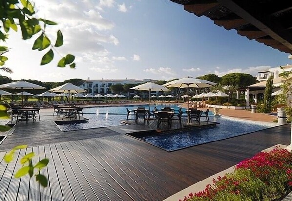 Zona de piscina con sillas de descanso y sombrillas, rodeada por una terraza de madera y una piscina.