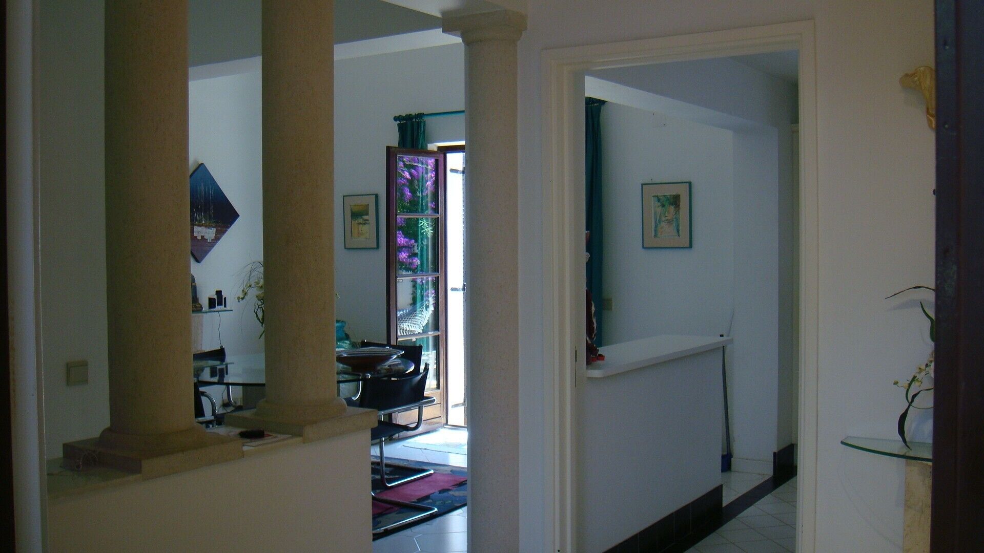 A hallway with a glass door leading to a dining area, a framed picture on the wall, and a small shelf with a plant.