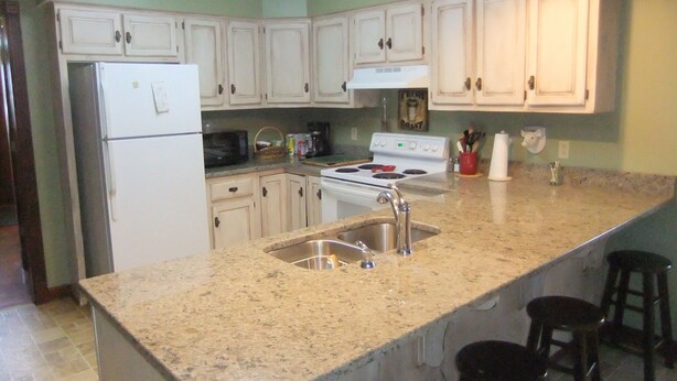 A kitchen with white cabinets, a granite countertop, a stainless steel sink, a stove, and a refrigerator. There are three stools at the counter.