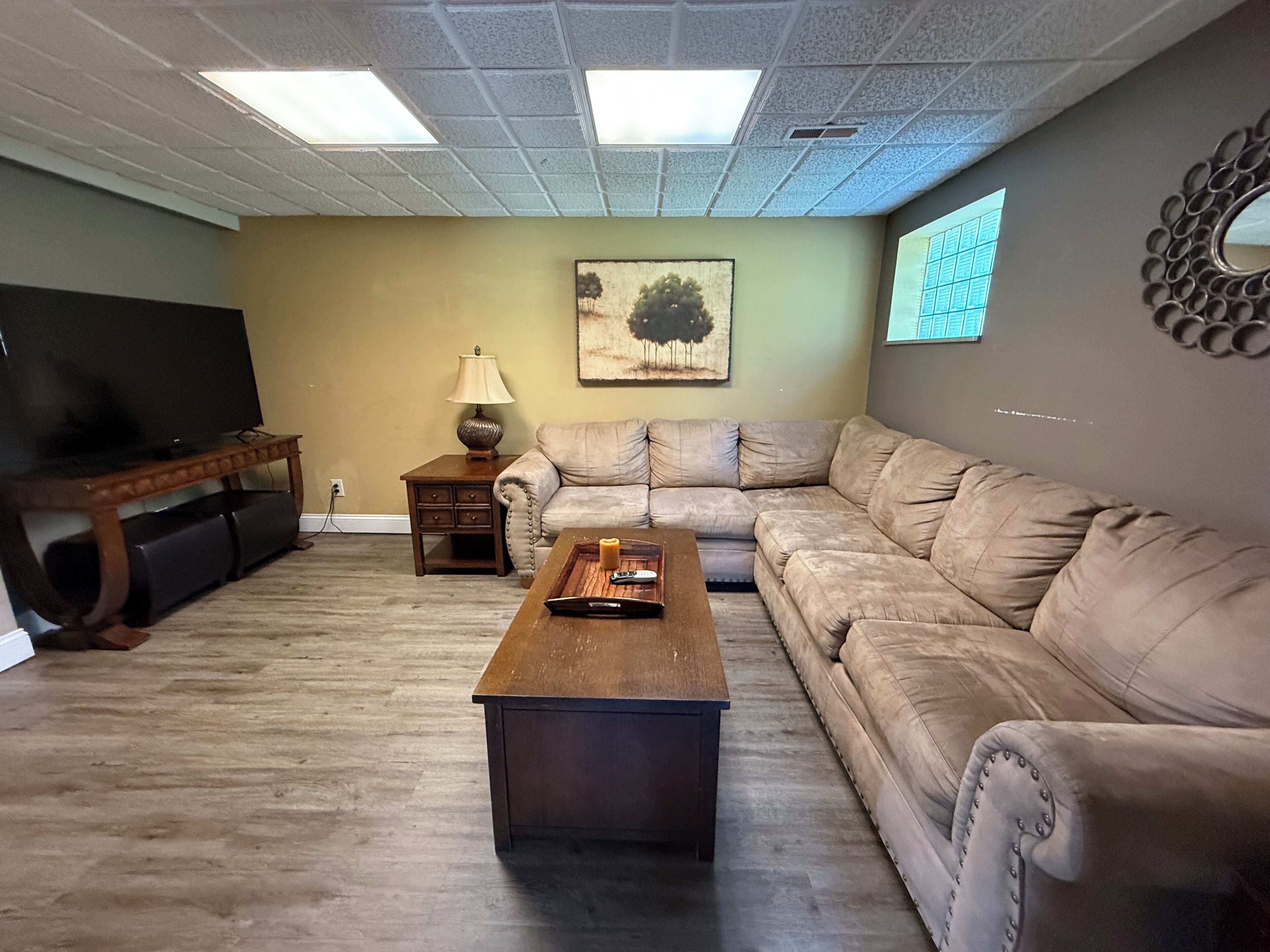 A living room with a flat-screen TV, a wooden coffee table, a beige sofa, a small side table with a lamp, and a painting on the wall.