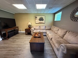 A living room with a flat-screen TV, a wooden coffee table, a beige sofa, a small side table with a lamp, and a painting on the wall.