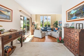 A spacious living room with a large window, a sofa set, a wooden cabinet, and a desk with a plant. - Includes beach gear! Waikoloa Colony Villas 1202 (Waikoloa Beach Resort)