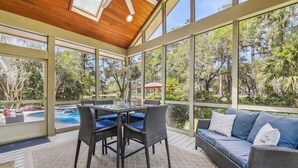 A covered outdoor area with a pool, wicker furniture, and a view of the surrounding trees. - Stunning Lagoon & Harbour Town Golf Views! 2 Master Suites! (Hilton Head Island)