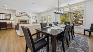 A modern dining area with a wooden table, black chairs, and a chandelier. There is a fireplace with a mantel, framed artwork, and a large window with a view of trees. - Stunning Lagoon & Harbour Town Golf Views! 2 Master Suites! (Hilton Head Island)