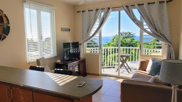 A room with a kitchen area, a dining table, a sofa, a TV, and a balcony with a view of the sea. - WaveView - Rincon Apartment Rentals:  Puntas 1  BR Condo w/ Ocean View (Puntas Rincon)