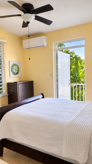 A bedroom with a bed, a ceiling fan, an air conditioning unit, a dresser, and a door leading to a balcony with greenery.
