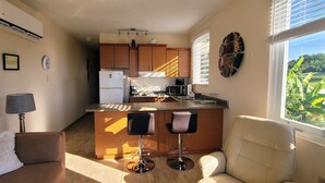 A compact living space with a kitchenette, a dining area with two bar stools, a sofa, and a window with a view of greenery. - WaveView - Rincon Apartment Rentals:  Puntas 1  BR Condo w/ Ocean View (Puntas Rincon)