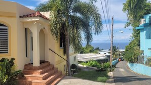 A residential street with a yellow house featuring a tiled roof, a staircase, and a small garden. - WaveView - Rincon Apartment Rentals:  Puntas 1  BR Condo w/ Ocean View (Puntas Rincon)