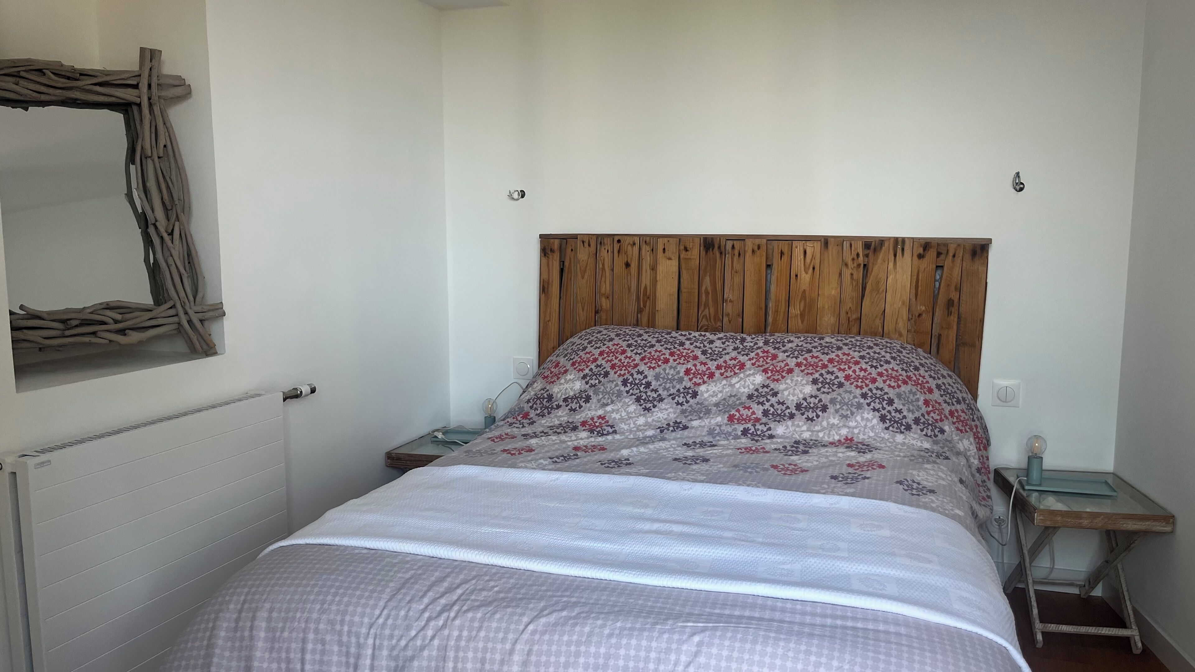 A bedroom with a wooden headboard, a patterned bedspread, a bedside table with a lamp, and a wall-mounted mirror.