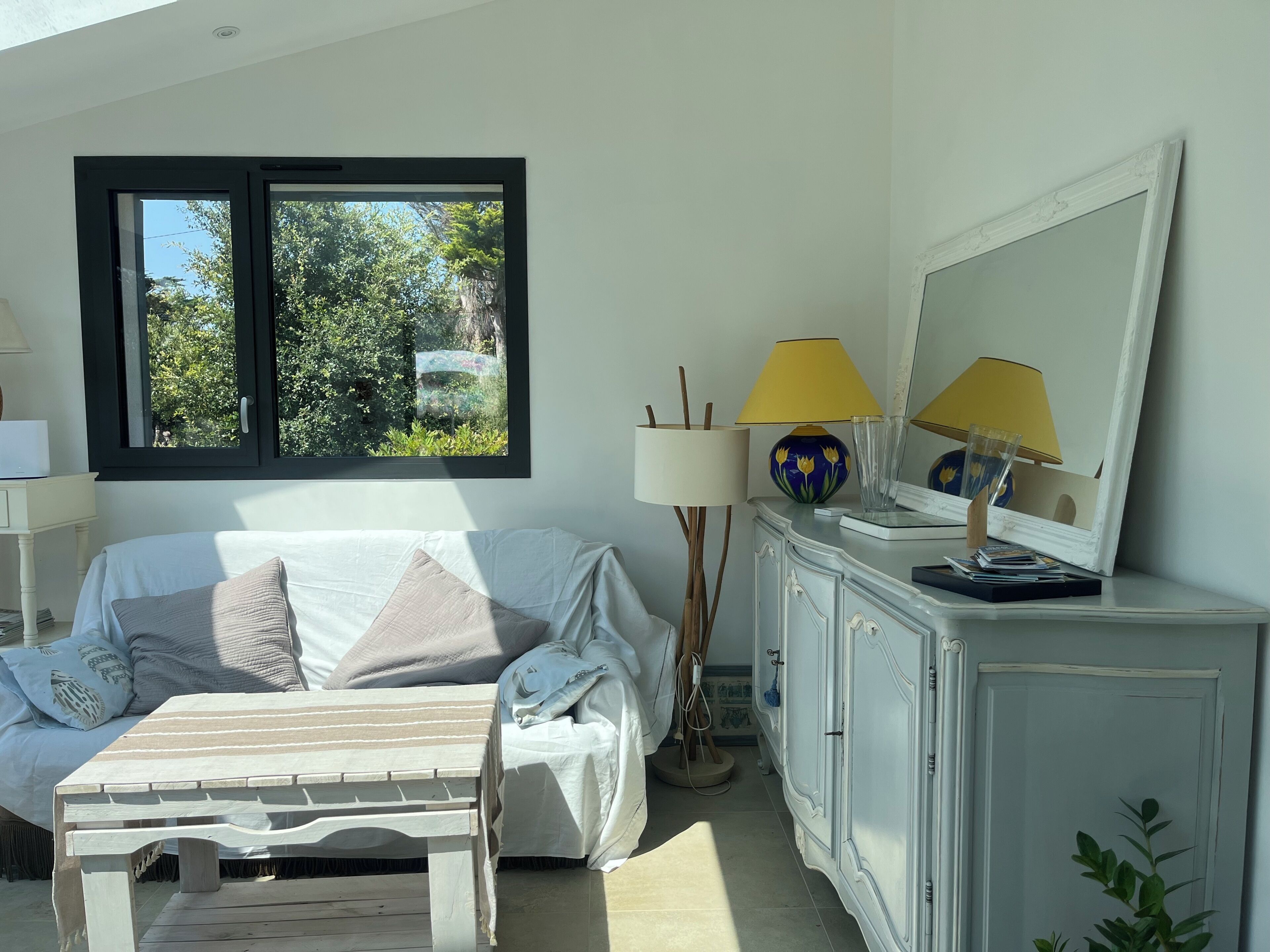 Una habitación con cama, mesita de noche, lámpara, espejo y ventana con vistas al verdor.