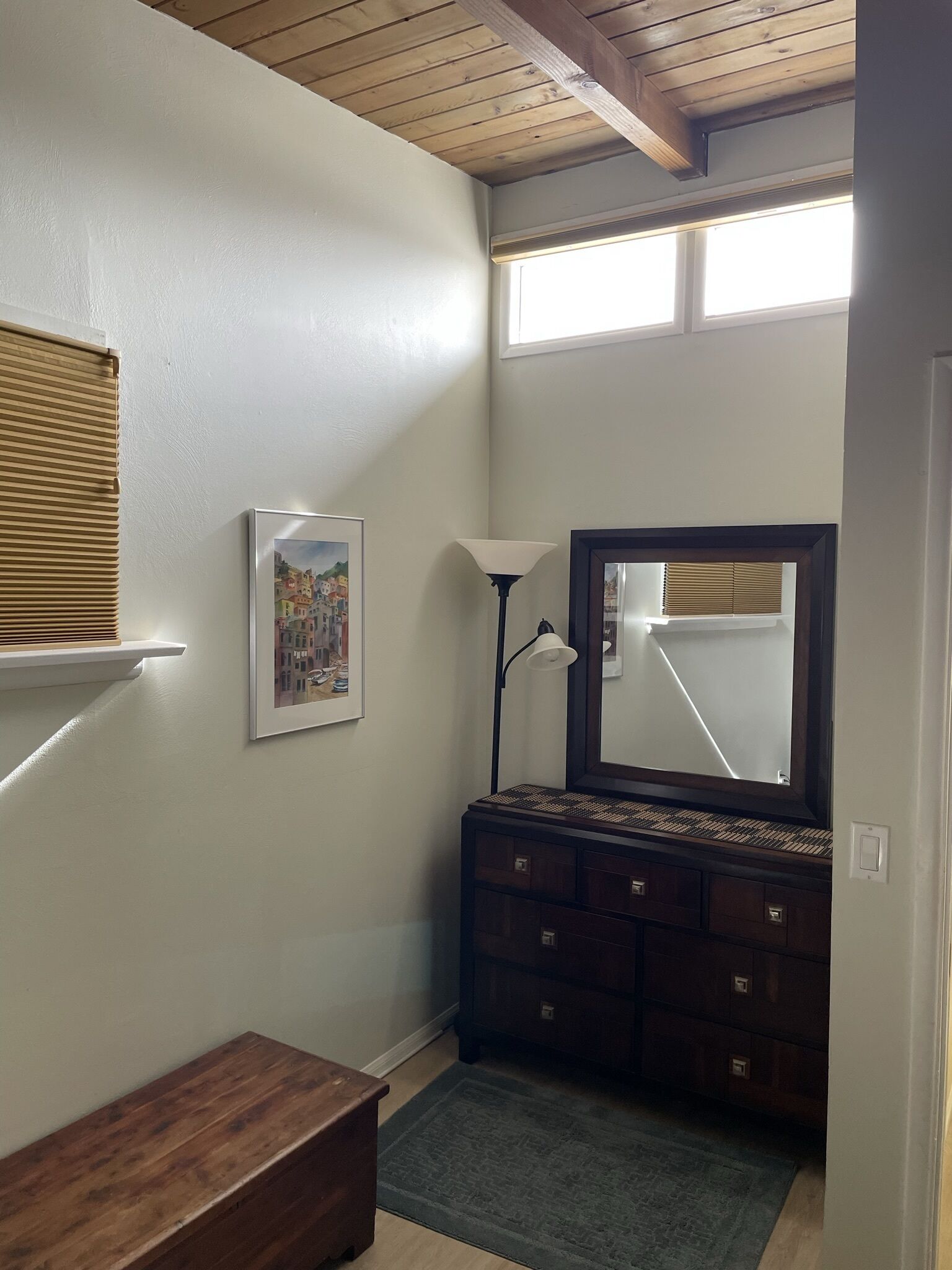 Una habitación con techo de madera, un aparador con varios cajones, un cuadro enmarcado en la pared, una lámpara de pie y una ventana con persianas.