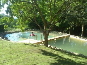 Zona de natación al aire libre bordeada de árboles, con un muelle de madera y una persona en un flotador rojo.