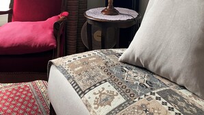Een hotelkamer met een rode fauteuil, een ronde tafel met een lamp, een vloerkleed met dessin, en een bed met een wit kussen.