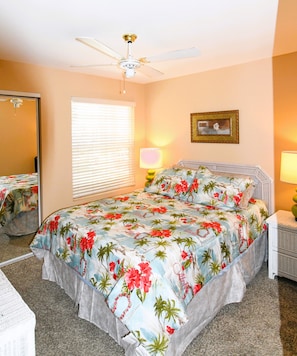 A bedroom with a bed, a ceiling fan, a window with blinds, a bedside lamp, a mirror, and a framed picture on the wall.