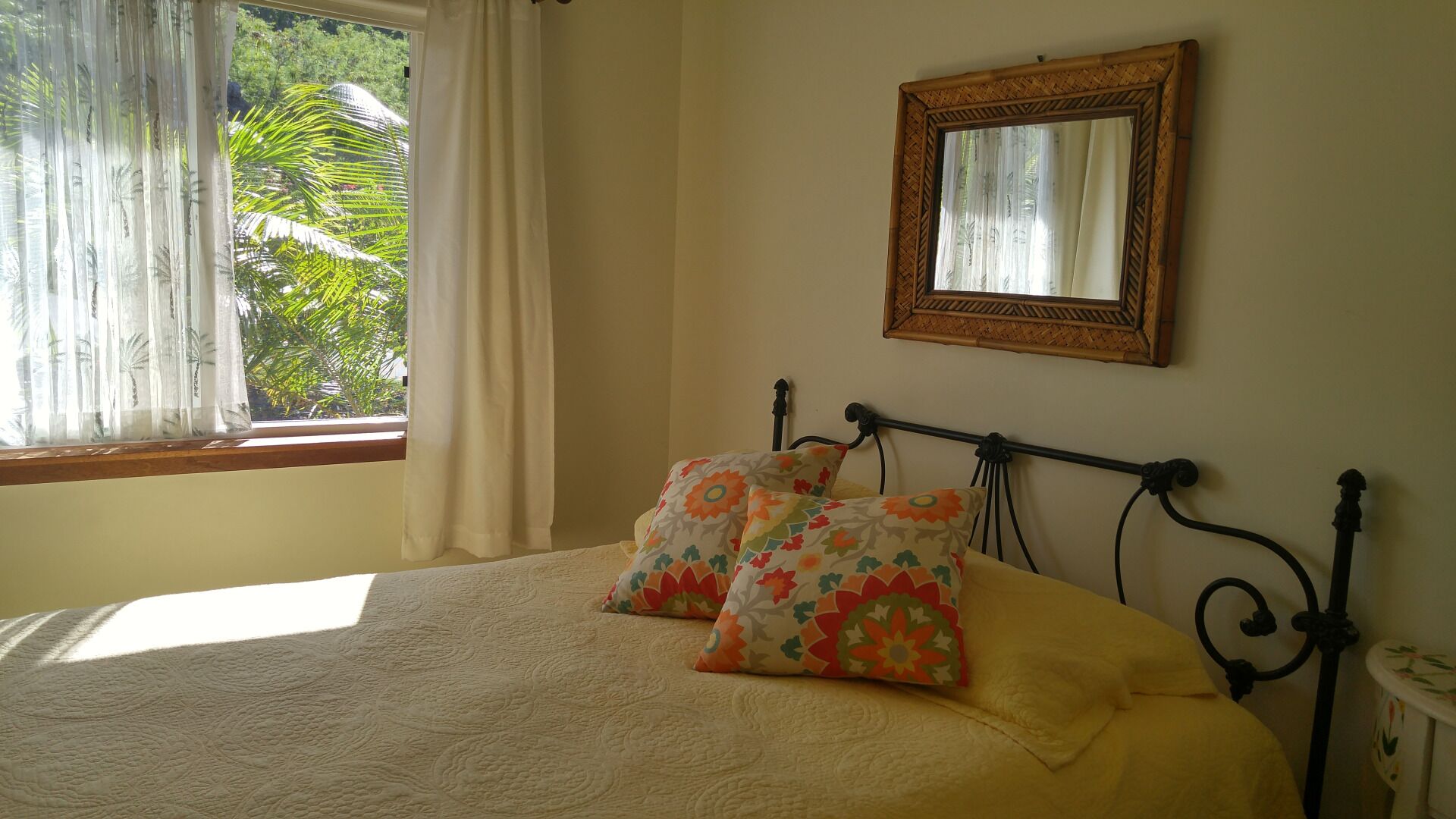 Una cama con colcha y almohadas estampadas con flores, una ventana con cortinas translúcidas y un espejo en la pared.