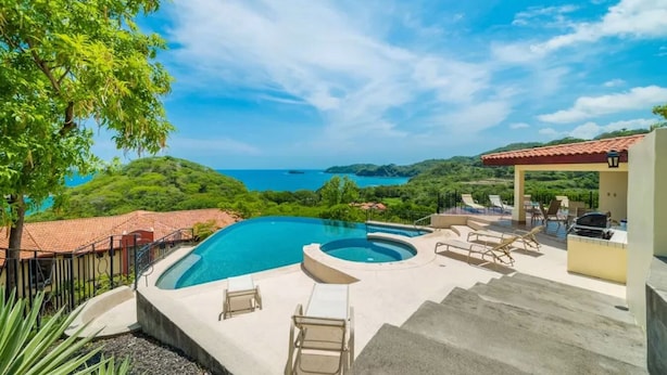 Une piscine avec des chaises longues, un jacuzzi et une vue sur l’océan et les collines.