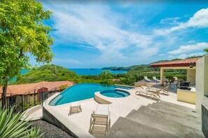 Une piscine avec des chaises longues, un jacuzzi et une vue sur l’océan et les collines.