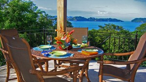 Une table ronde entourée de chaises, dressée pour un repas, offrant une vue panoramique sur un plan d’eau et des collines.