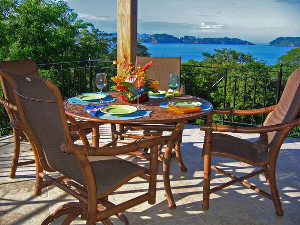 Une table ronde entourée de chaises, dressée pour un repas, offrant une vue panoramique sur un plan d’eau et des collines.