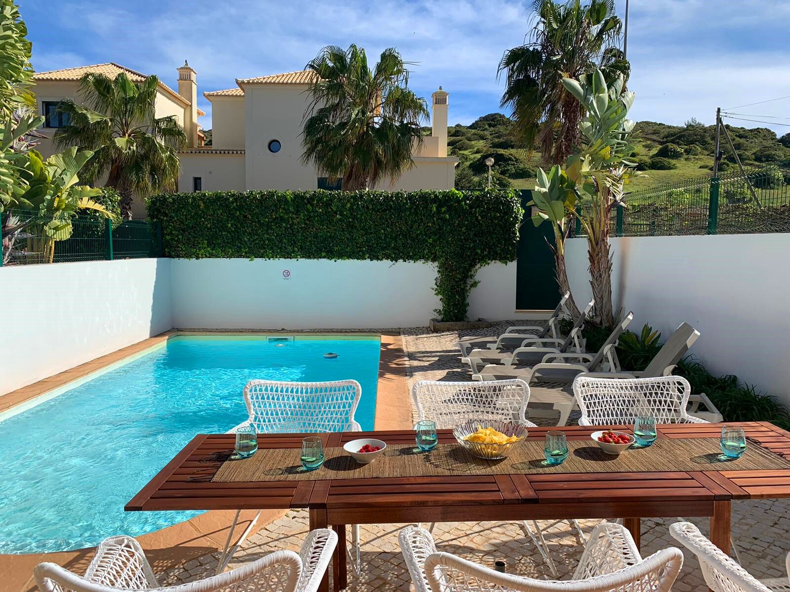 A poolside area with a wooden table, white wicker chairs, and a view of a house with a tiled roof and a green hedge.